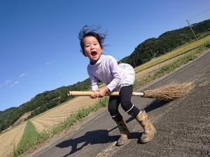 青い空と緑の木々、広大な田んぼを背景に魔女のようにホウキに乗ってるかわいらしい女の子の写真