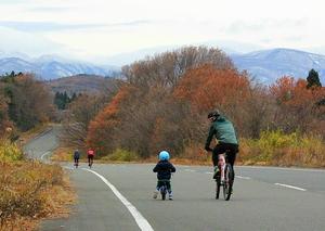 遠くに山が見え紅葉している木々が並ぶ道路を親子でサイクリングしている様子の写真