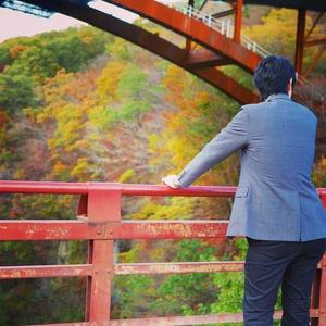 スーツを着た男性が山の中にある赤い陸橋に立ち黄色やオレンジ色に紅葉する木々を眺めてる様子の写真
